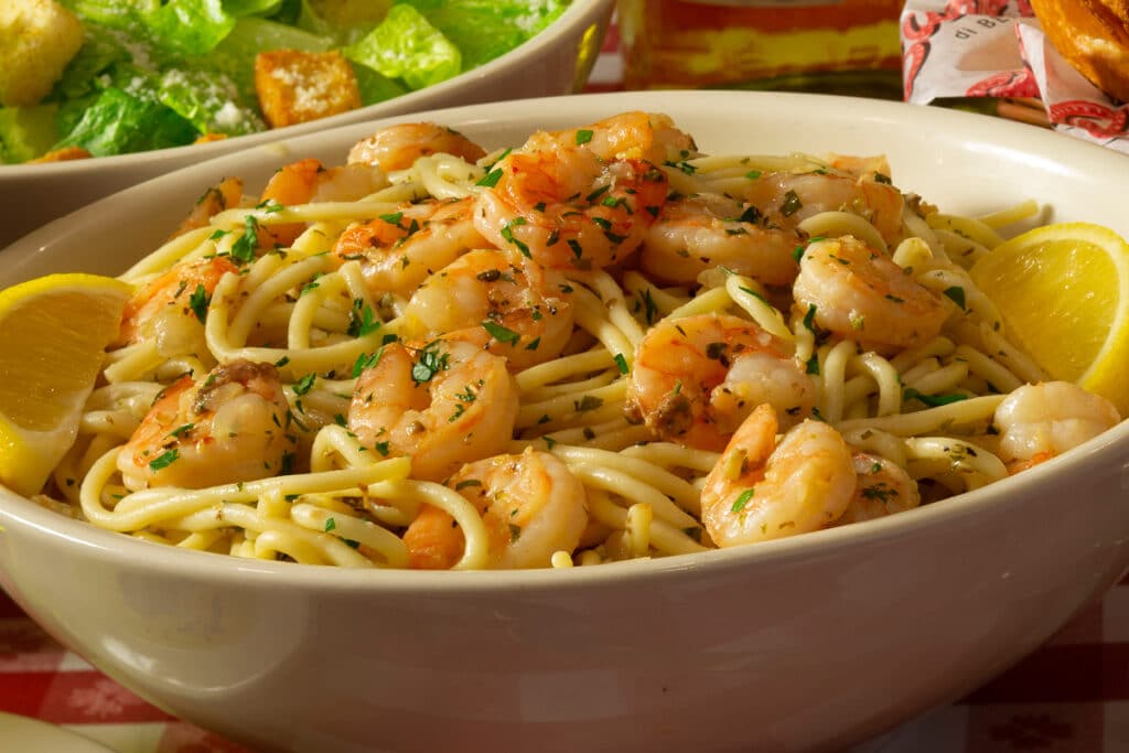 Closeup of Buca di Beppo Shrimp Scampi in a bowl with salad and garlic bread in the background